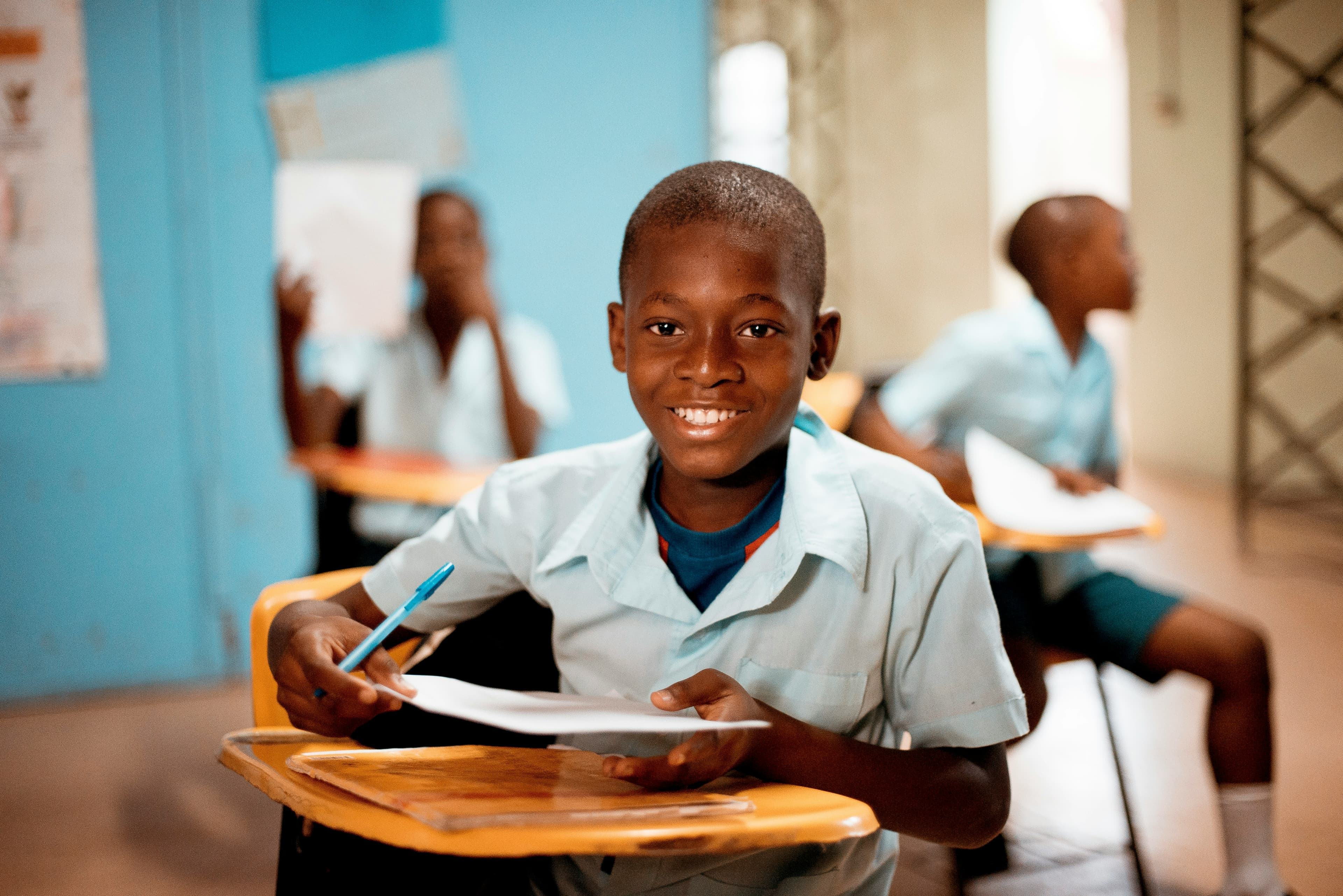 Student at a desk in a Ghanaian classroom, smiling at the camera